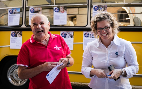 Ein Besucher im roten Hemd lacht mit einer Botschafterin; im Hintergrund steht das Postauto Meet the Ambassador