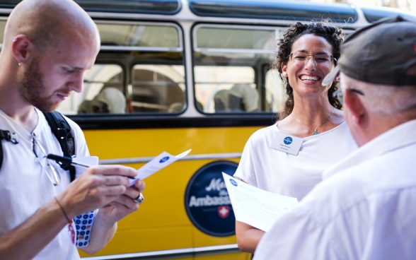 A dark-haired ambassador talks to two visitors; in the background is the bus Meet the Ambassador