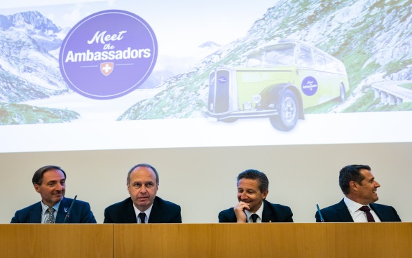 Vier Botschafter auf dem Podium der Aula, im Hintergrund ein Foto des Postautos Meet the Ambassador