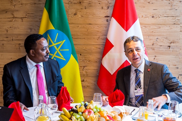 An einem reich gedeckten Tisch sitzt Bundesrat Cassis in Gespräch vertieft mit dem äthiopischen Aussenminister Gebeyehu. Im Hintergrund sind die Flaggen der Schweiz und Äthiopiens zu sehen.