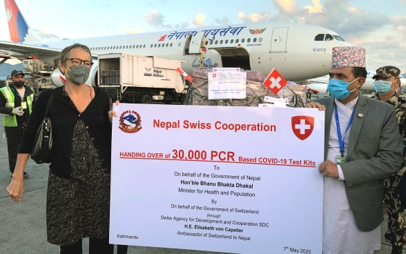 The Swiss ambassador to Nepal and the Nepalese Minister for Health and Population standing in front of an airplane and holding up a poster announcing the handover of 30,000 COVID-19 test kits.