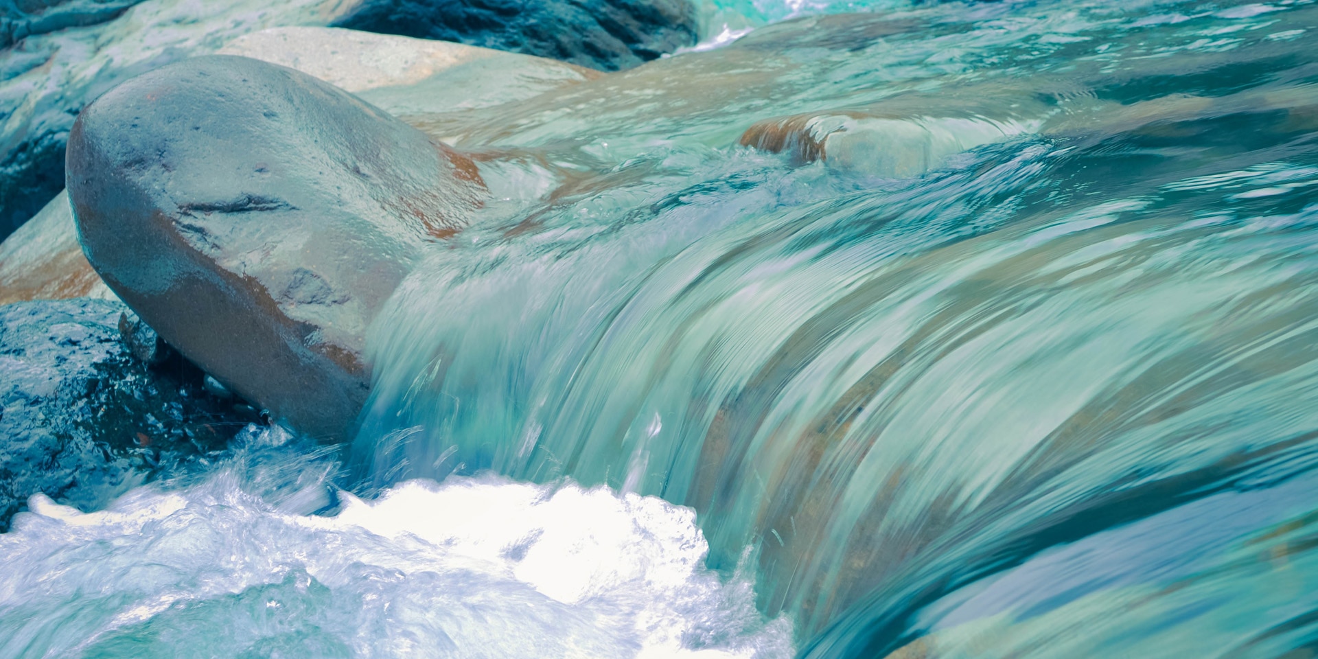 Das blaue Wasser eines Baches plätschert über steinigen Untergrund.