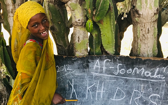 Ein Mädchen steht vor einer Wandtafel im Freien und schaut in die Kamera (Bild aufgenommen in Äthiopien). 