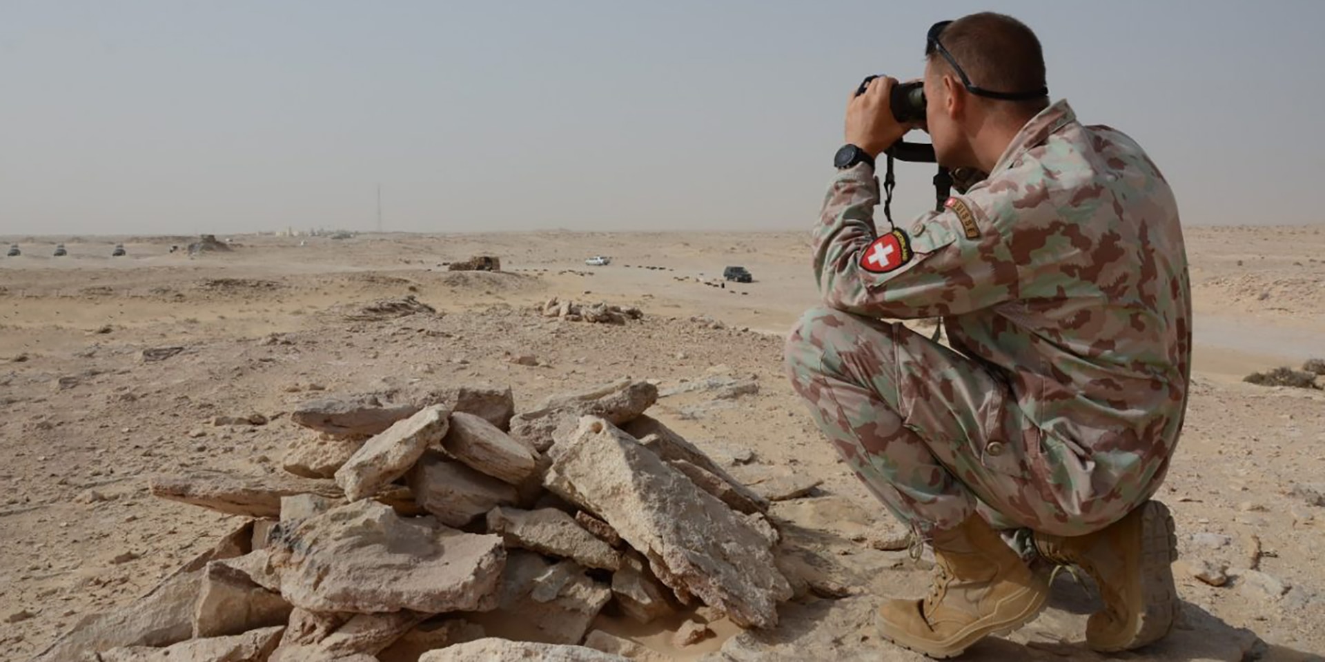 Ein Soldat der Schweizer Armee kniet im Sand und schaut mit dem Fernglas in die Wüste.