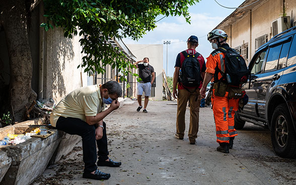 Un uomo è seduto disperato sul ciglio della strada con la testa tra le mani. Gli esperti dell’Aiuto umanitario della Confederazione gli passano accanto. 