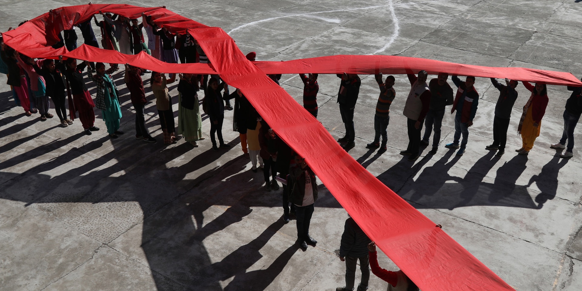 Viele Personen heben ein langes rotes Band in Form einer Schlaufe über ihren Köpfen. Sie ist weltweit ein Symbol für den Kampf gegen Aids.