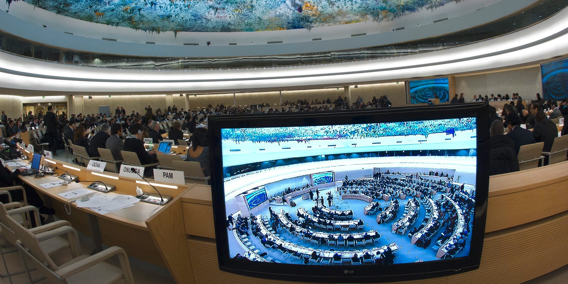 The Human Rights and Alliance of Civilizations Room at the United Nations in Geneva, where the Human Rights Council convenes. In the foreground, a screen showing footage of the conference room.
