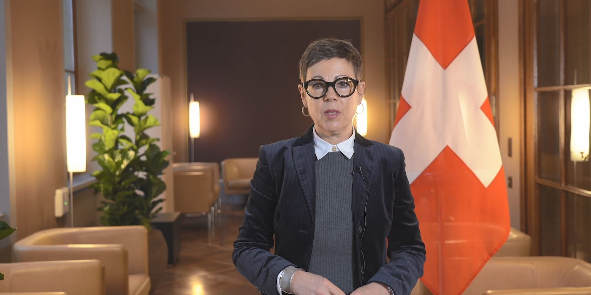 La secrétaire d'État du DFAE Krystyna Marty Lang se tient dans une salle lambrissée et parle devant une caméra. On peut voir le drapeau de la Suisse en arrière-plan.