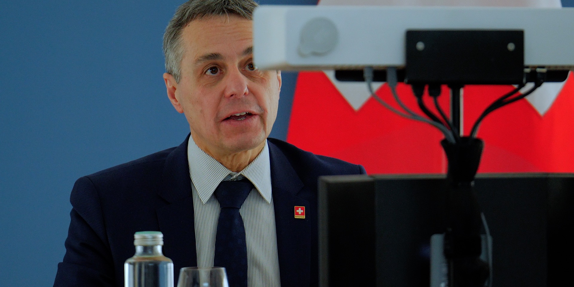 Il consigliere federale Ignazio Cassis davanti a una telecamera durante una conferenza virtuale, la bandiera svizzera in sottofondo.