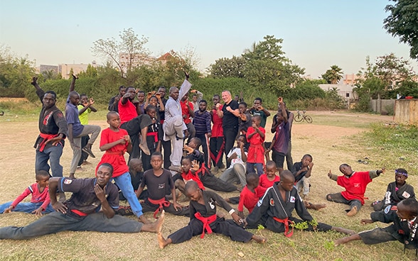 Carl Emery in simple black outfit poses with about forty children from Bamako in black judogi and red hinge.