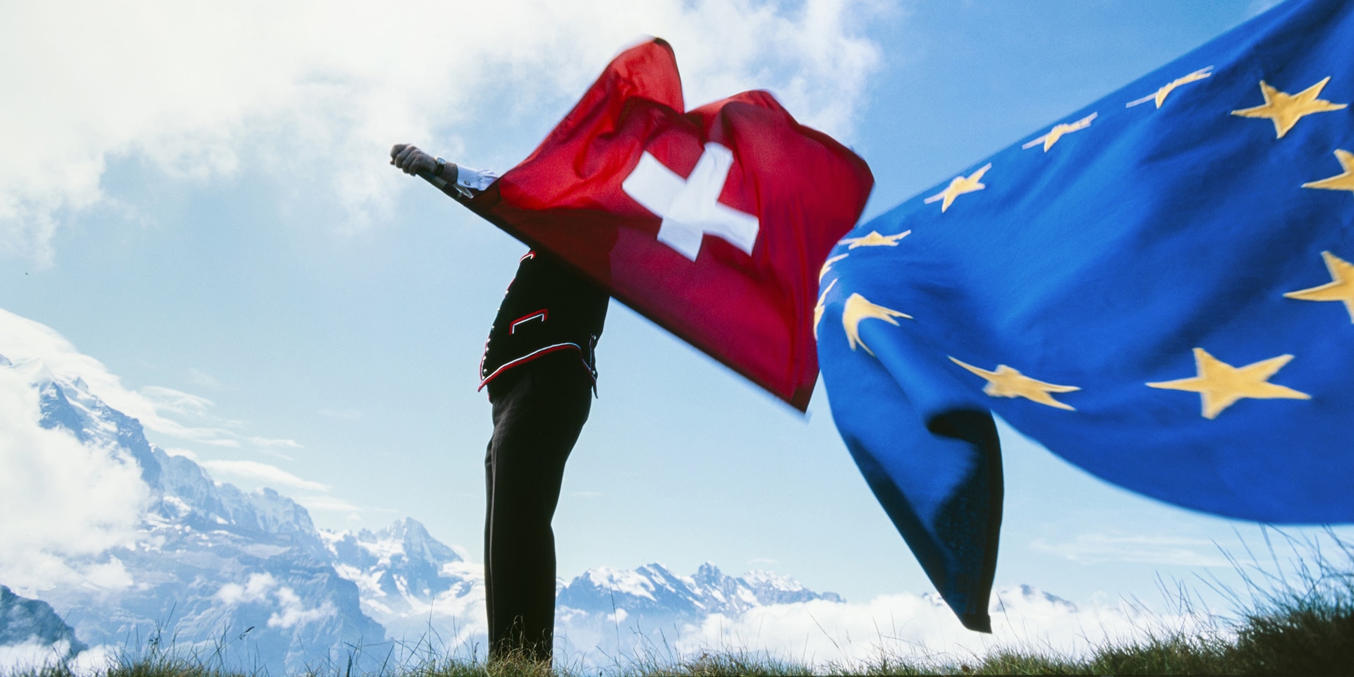 A flag-thrower waving the Swiss flag in one hand and the EU flag in the other against the backdrop of the Bernese Alps.