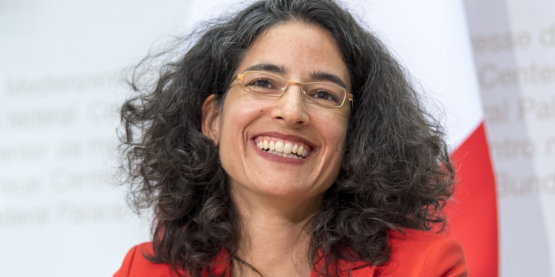 Portrait of Corinne Cicéron Bühler, smiling with the Swiss flag in the background.