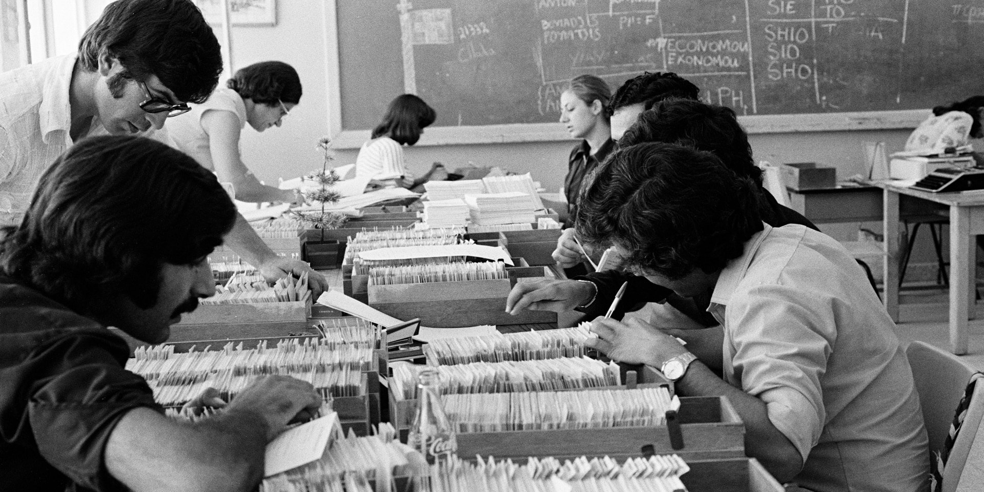 The picture, black and white, shows an office space in 1974. The desks are covered with index boxes filled with index cards. Eight people, concentrated, are working hard.  