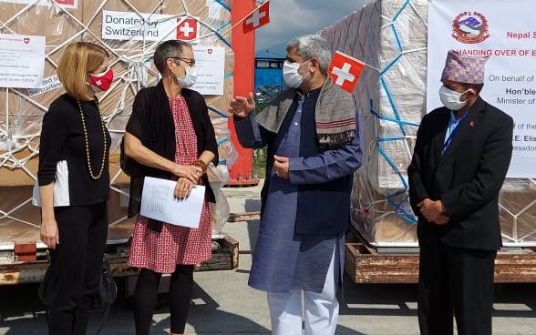 Four people, including Swiss Ambassador to Nepal Elisabeth von Capeller, stand on an airfield in Kathmandu in front of a pallet of relief supplies.