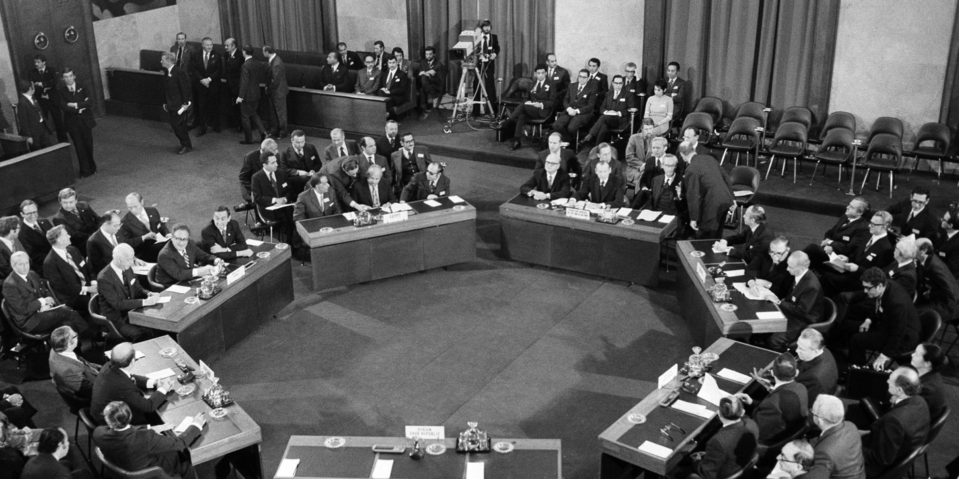 Photographie en noir et blanc: sept tables sont disposées en cercle dans une salle de conférence, avec des hommes assis à ces tables.
