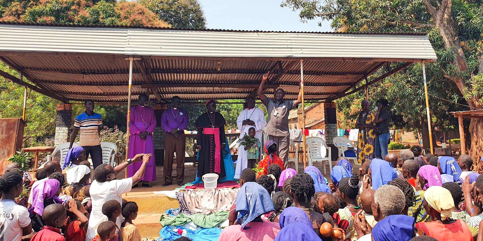 Five men are standing on a podium, recognisable by their clothing as representatives of different religious communities. One of them is speaking to an audience in the foreground of the photo.