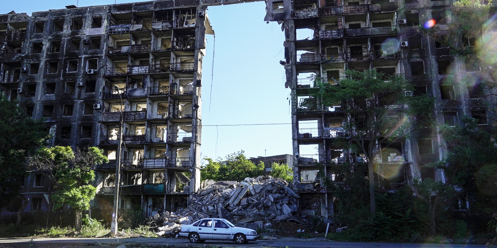 Un’auto bianca è ferma davanti a un edificio civile distrutto, attraverso il cui scheletro si scorge il cielo blu.