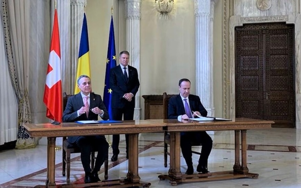 Le président de la Confédération Cassis et le ministre roumain des Finances Adrian Caciu sont assis côte à côte à une table et signent tous deux un accord.