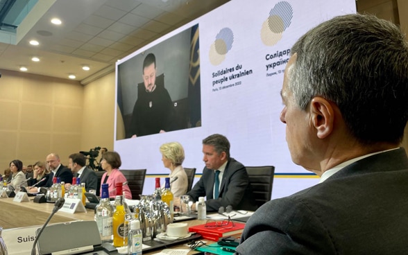 Bundesrat Cassis sitzt am Tisch und hört dem ukrainischen Präsidenten Selenskyj zu, der per Video auf einer Leinwand zu sehen ist.