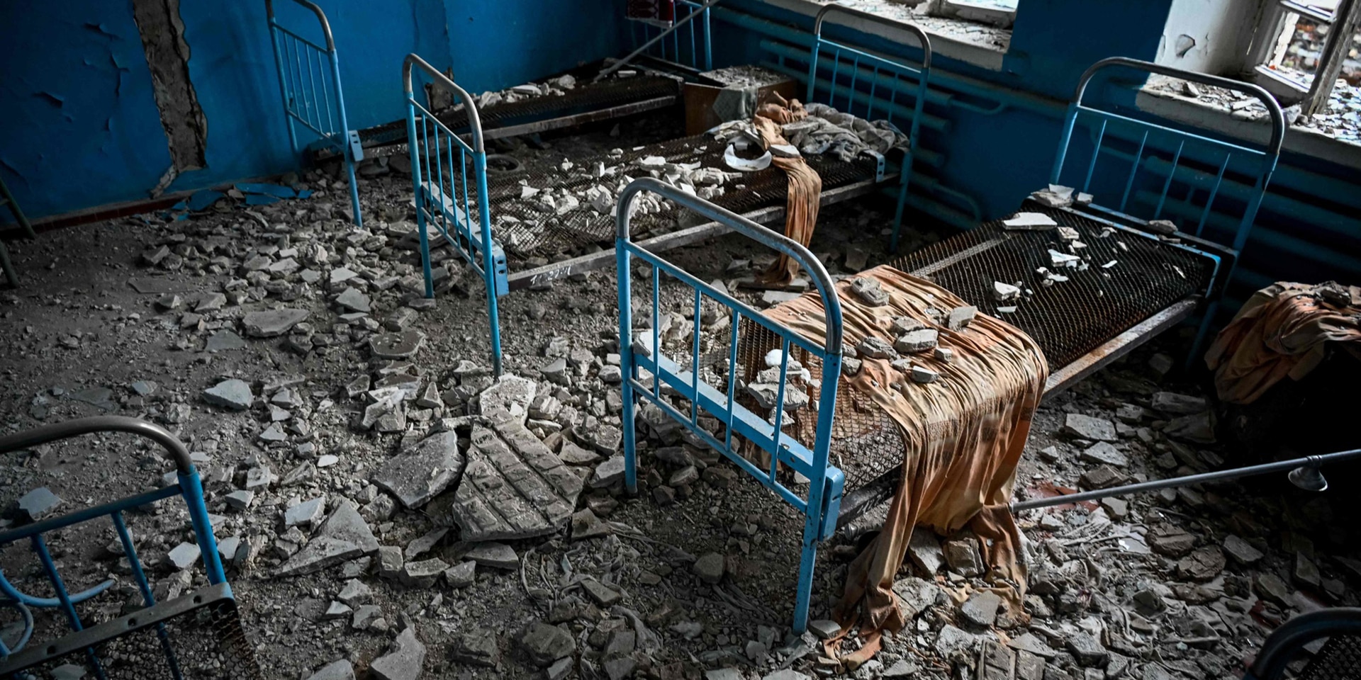 In einem Zimmer, dessen Fenster und Wände beschädigt ist, stehen vier Bettgestelle. Der Boden ist mit Trümmern der Wände übersät.