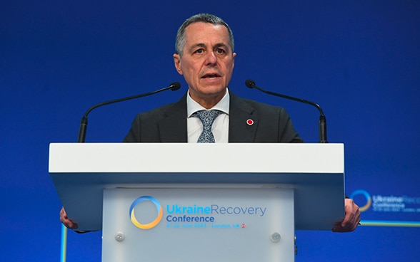 Le conseiller fédéral Ignazio Cassis se tient à un pupitre et prend la parole. Sur le devant du pupitre, on peut voir le logo de l’Ukraine Recovery Conference 2023.