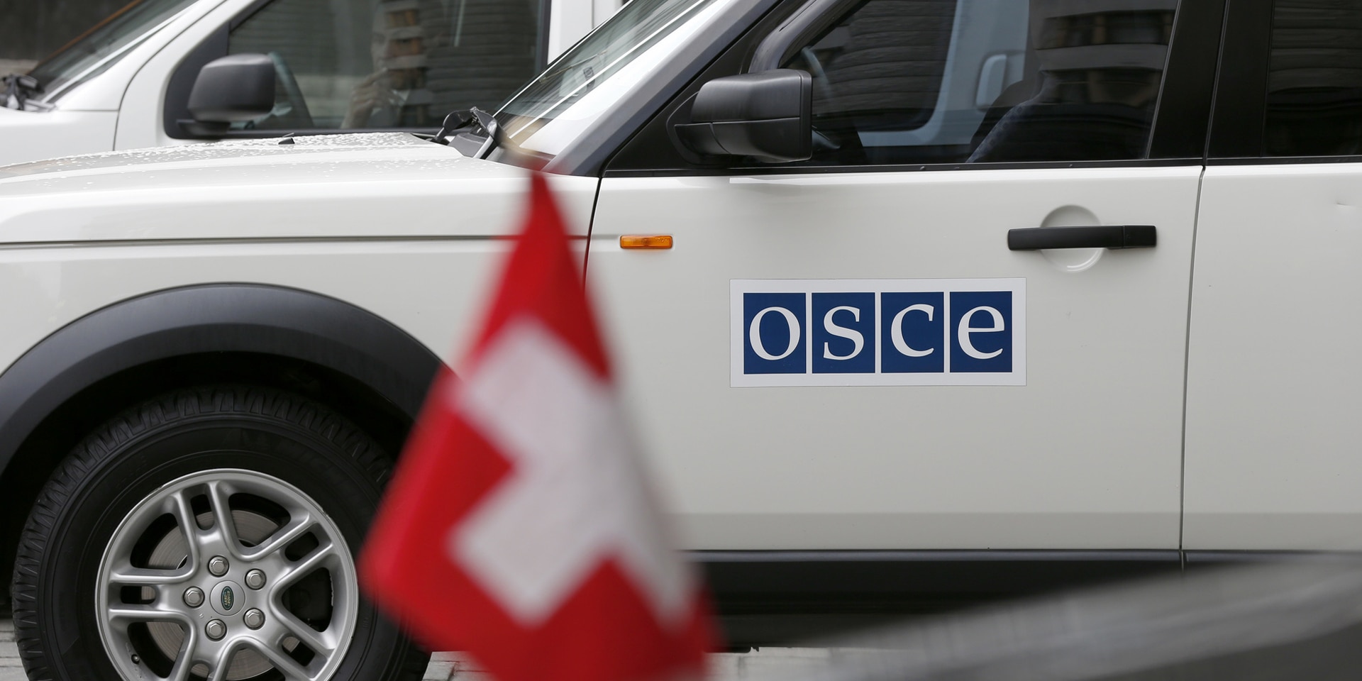 A white off-road vehicle labelled "OSCE" stands in front of a black car bearing the Swiss flag.