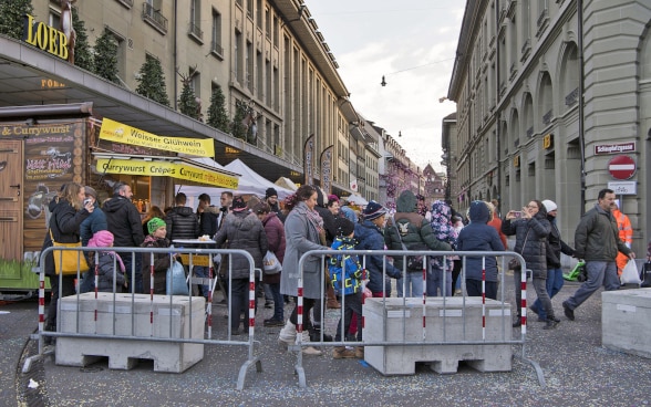 Des blocs de béton bloquent une rue de la capitale suisse, Berne. Derrière, un marché accueille de nombreuses personnes.