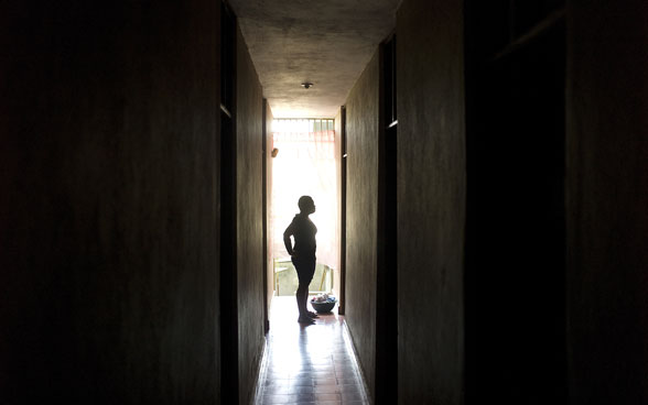 A woman appears against the light at the end of a corridor.