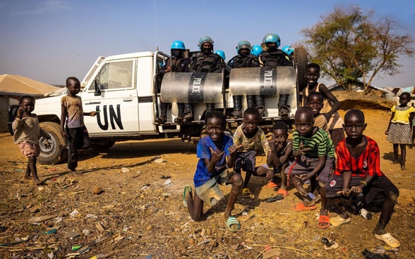 Eight UN police officers sit on the back of a white pick-up. In the foreground, African children pose for the photo.