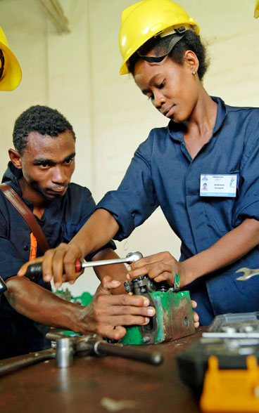 A trainer, wearing a yellow helmet, helps an apprentice in a workshop.