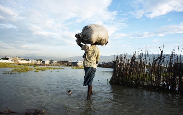 A woman carrying a sack on her head wades through water up to her ankles towards a group of houses. 