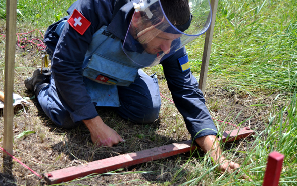 A man wearing protective clothing and a helmet kneels on the ground to defuse a landmine.