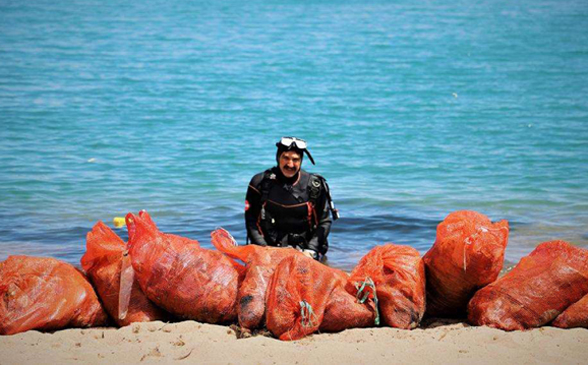 Un plongeur se tient dans l'eau au bord de la mer devant de nombreux sacs orange posés sur le sable.