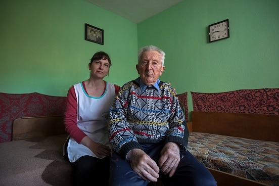 An elderly man sitting on a sofa in his house with a caregiver.