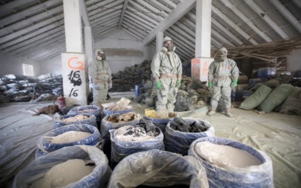 Three men in protective clothing next to containers of toxic substances.