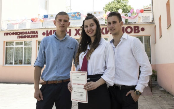 Drei junge Personen, zwei Männer und eine Frau, welche ein Zertifikat hält, stehen vor einer Schule.